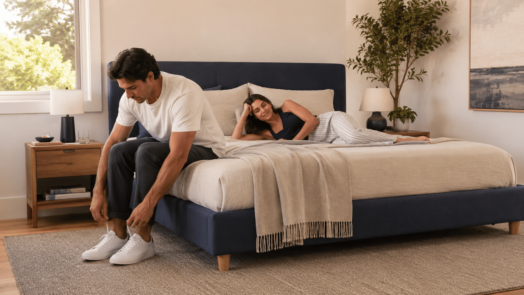 A realistic modern Australian bedroom showing one person sitting naturally on the mattress edge while another lies near the opposite side.